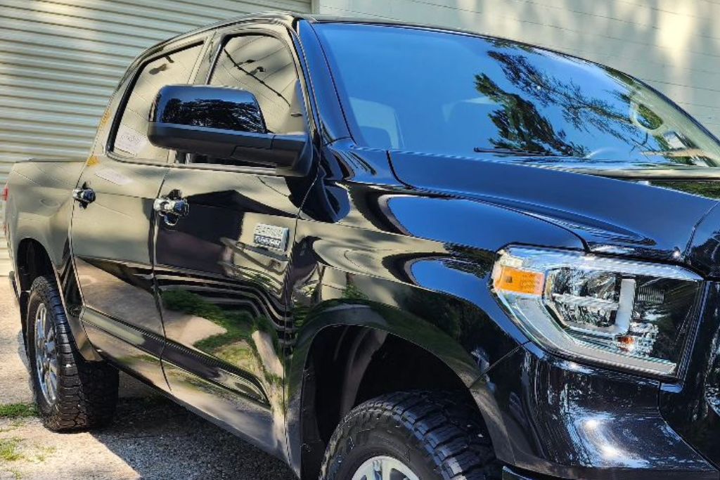 The image shows a black pickup truck, with dark-tinted windows, parked in an outdoor setting. The truck has a highly reflective, polished surface that mirrors its surroundings, including trees and nearby structures. The vehicle appears clean and well-maintained, with clear, detailed headlights and black side mirrors. The background features a garage door and greenery, adding to the contrast between the sleek truck and its environment.