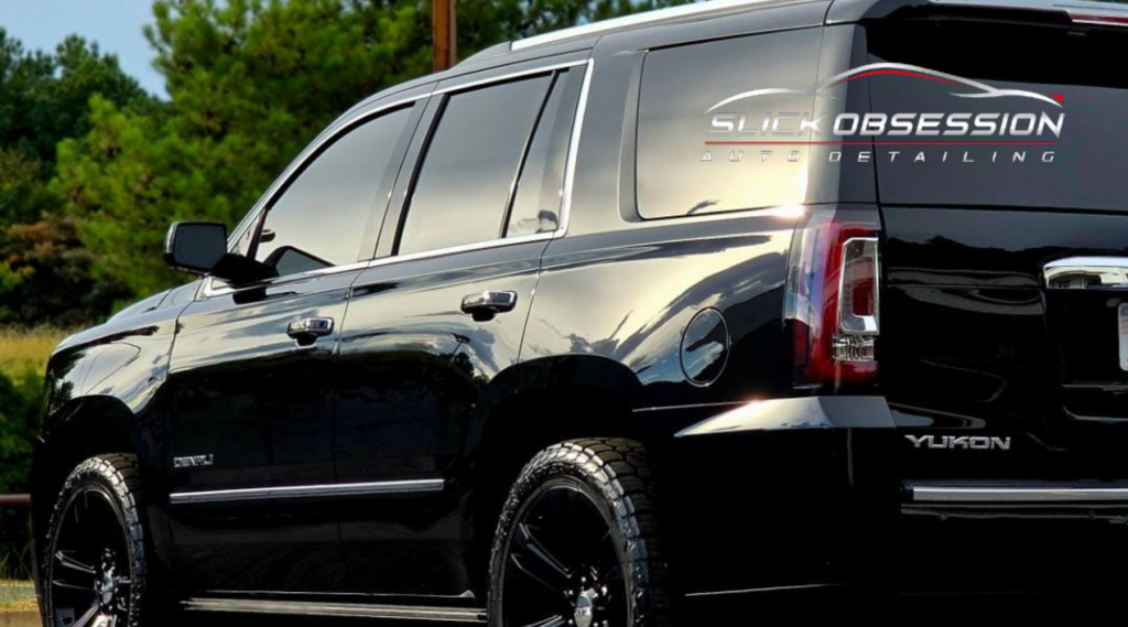 image shows a glossy black SUV, specifically a GMC Yukon Denali, with tinted windows and reflective detailing. The vehicle has "Slick Obsession Auto Detailing" branded on the rear window, showcasing a professional finish likely after detailing or tinting services. The background features greenery, Window Tint Installation in Birmingham, AL How Quality Affects Your Comfort and Safety - suggesting the vehicle is parked outdoors.