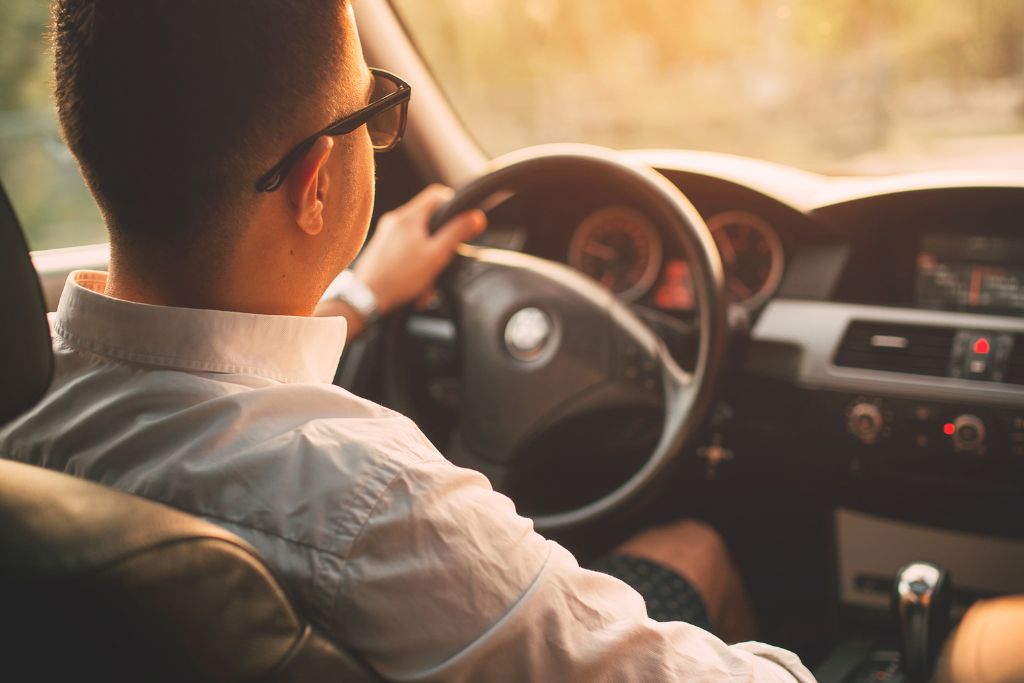 A person wearing sunglasses drives a car with sunlight streaming through the window, highlighting the comfortable interior. The scene emphasizes the benefit of tinted windows for enhanced comfort during sunny days.