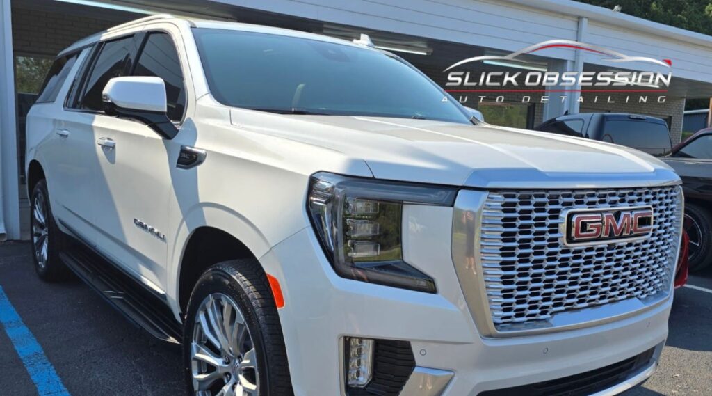 A freshly detailed white GMC Denali SUV with a sleek exterior finish, parked in front of a building featuring the "Slick Obsession Auto Detailing" logo. The vehicle’s chrome grille and large wheels reflect the meticulous detailing work.