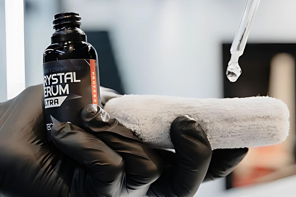 A gloved hand applying drops of Crystal Serum Ultra ceramic coating onto an applicator pad, preparing to coat a car's surface for protection.