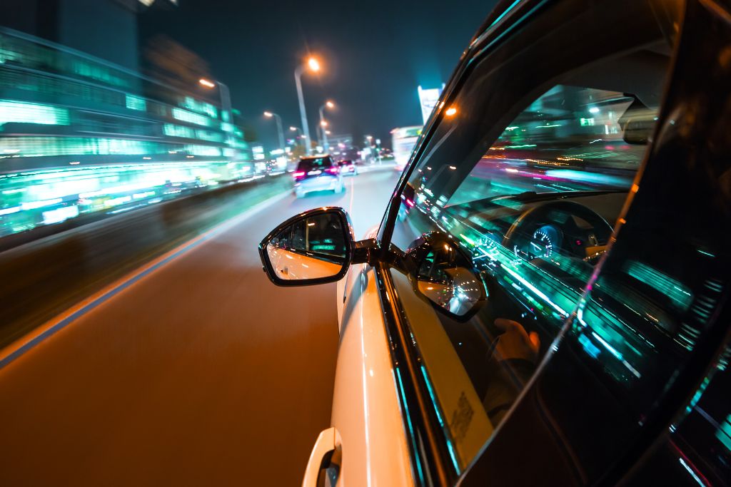 A car is driving down a brightly lit highway at night, with the lights of the city reflecting off the vehicle's windows. The image showcases the role of window tinting in reducing glare for better night driving.