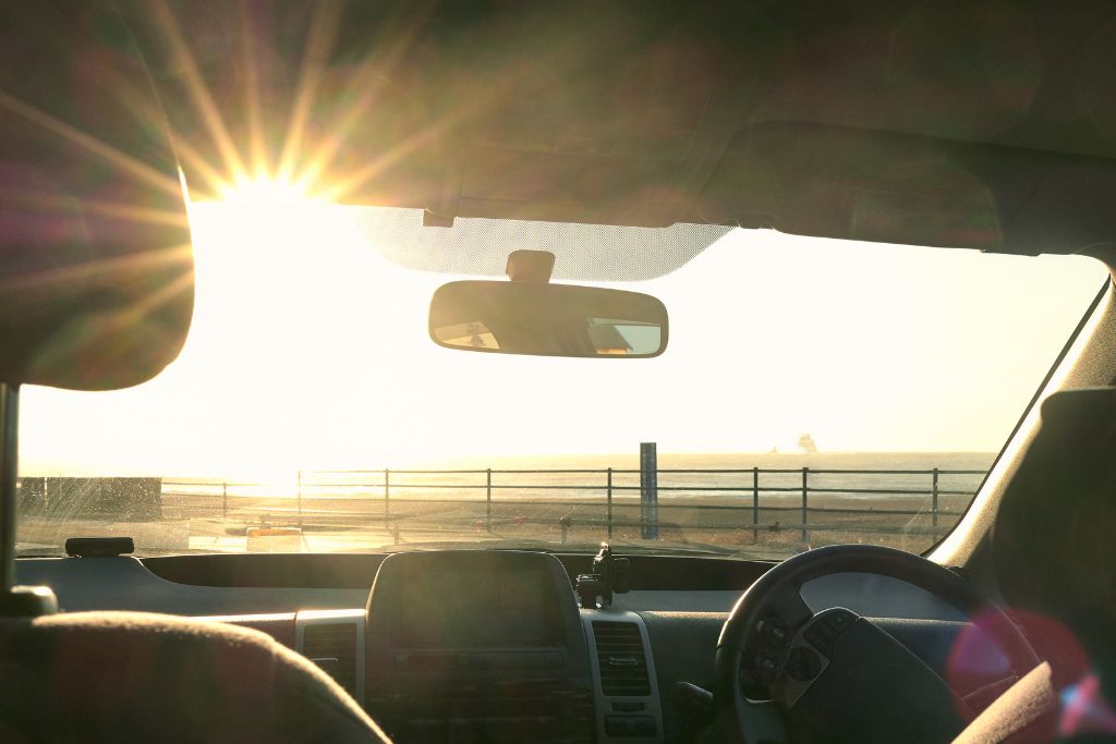 A car's interior with a view of the sun shining directly through the front windshield, creating a bright glow. The scene suggests the importance of tinted windows for skin protection and reducing UV exposure while driving.