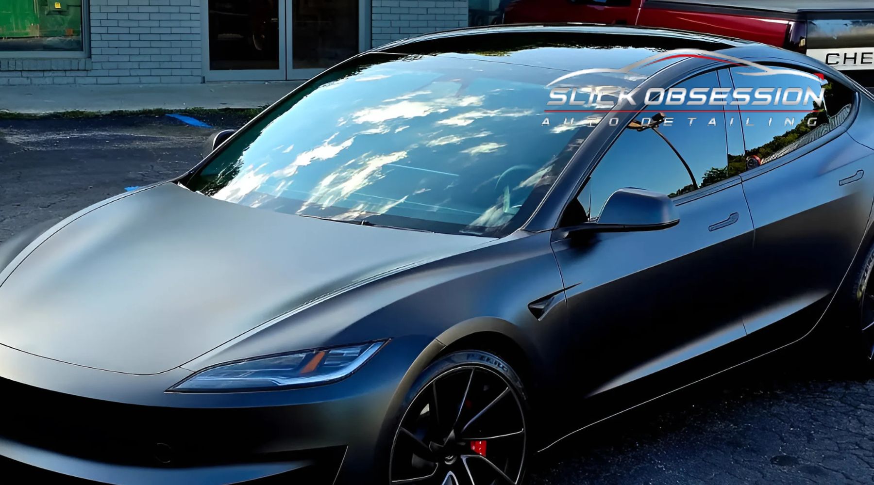 The image shows a sleek, dark-colored car with a glossy finish, featuring high-quality window tinting that reflects the blue sky and clouds. The logo "Slick Obsession Auto Detailing" is prominently displayed, suggesting professional car detailing services.