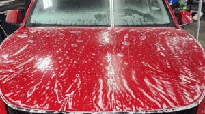 A red car's hood with paint protection film (PPF) being applied, covered in soapy water to aid in the installation process. The windshield is partially visible, and the bright indoor lig