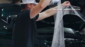 A technician wearing a white cap and black shirt applies a sheet of clear protective film to a car, with the logo "Slick Obsession Auto Detailing" visible in the background. The car's hood is open, indicating a professional detailing process in progress.