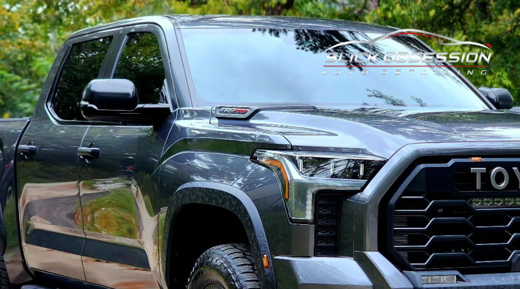 A sleek, black Toyota truck with tinted windows and a bold grille design, featuring a TRD Pro badge on the hood. The truck is parked outdoors, reflecting surrounding greenery and a road in its glossy finish, with the "Slick Obsession Auto Detailing" logo overlayed on the image.