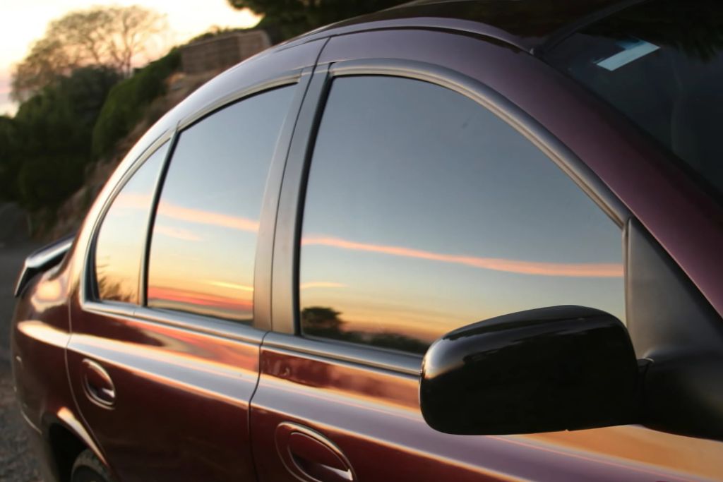 A side view of a car with dyed window film, reflecting a colorful sunset and surrounding landscape. The tinted windows provide a darker appearance for added privacy and style, ideal for those seeking an affordable aesthetic upgrade.