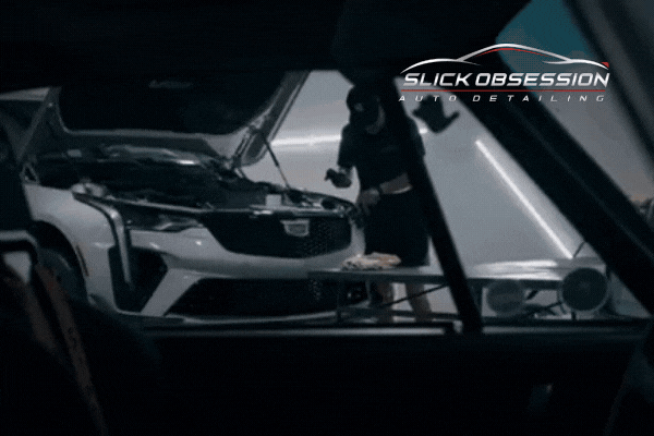 A professional detailer at Slick Obsession Auto Detailing applies a protective coating to a white Cadillac in a high-end detailing shop. The car's hood is open, and the technician works meticulously under bright studio lighting. The Slick Obsession Auto Detailing logo is displayed in the corner, emphasizing the brand’s premium services.