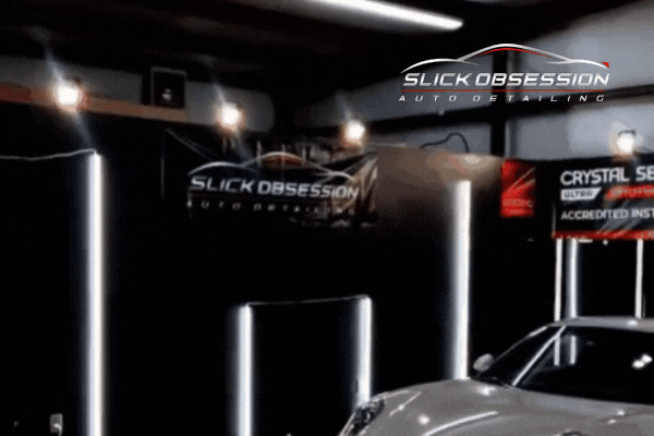 A sleek white sports car sits under bright studio lighting in a high-end detailing shop. The "Slick Obsession Auto Detailing" logo is displayed on banners in the background, emphasizing the professional environment.